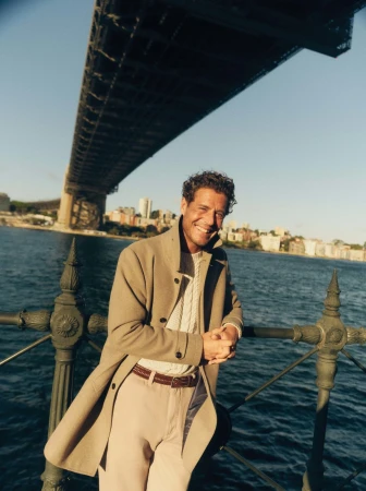 Adult man smiling and wearing a tan overcoat in an outdoor portrait with the Harbour Bridge and the harbor in the background.