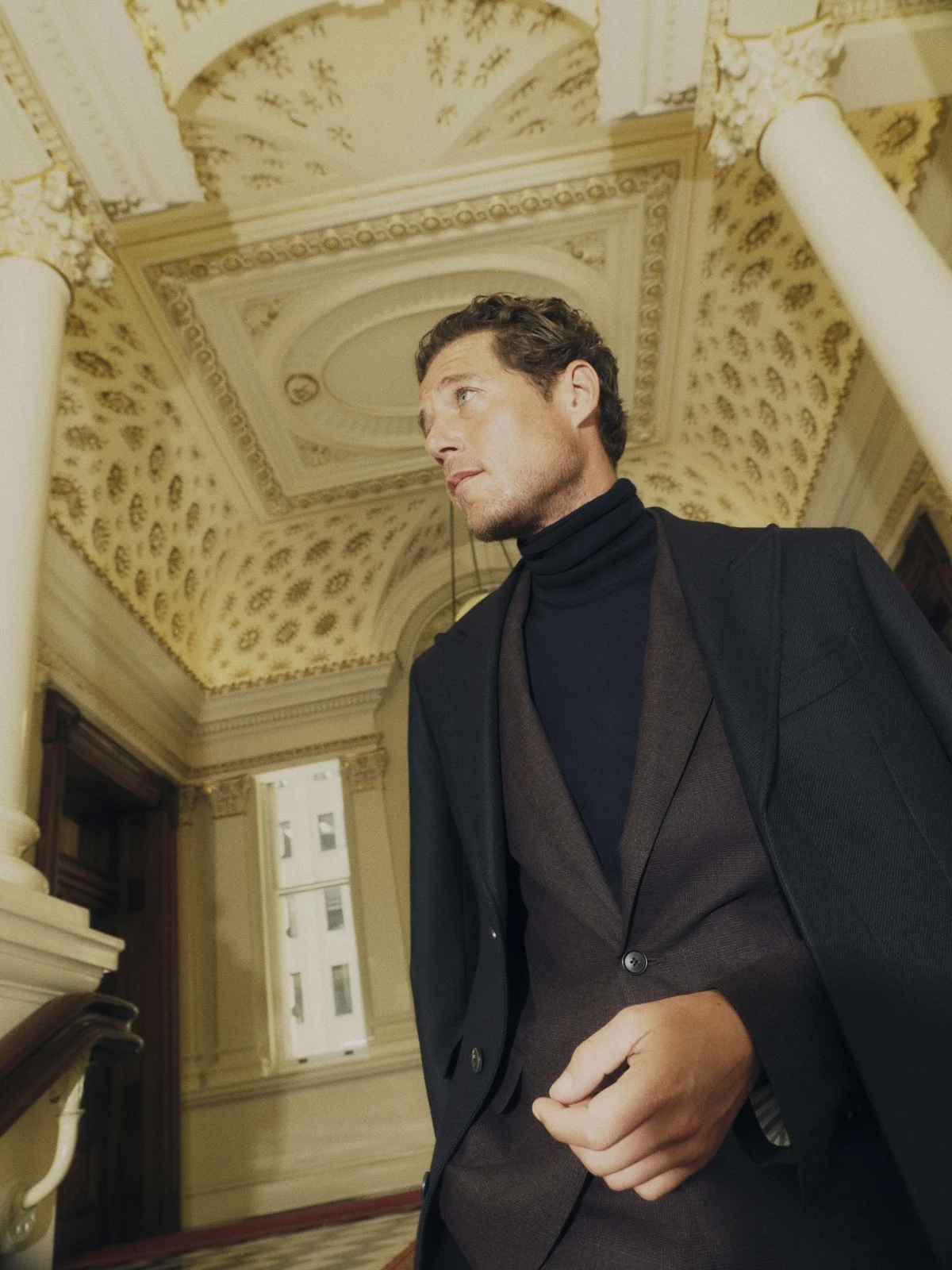 A man in a suit poses in a building with ornate ceilings, wearing a dark blue turtleneck, dark grey vest, and a dark overcoat in a formal setting.