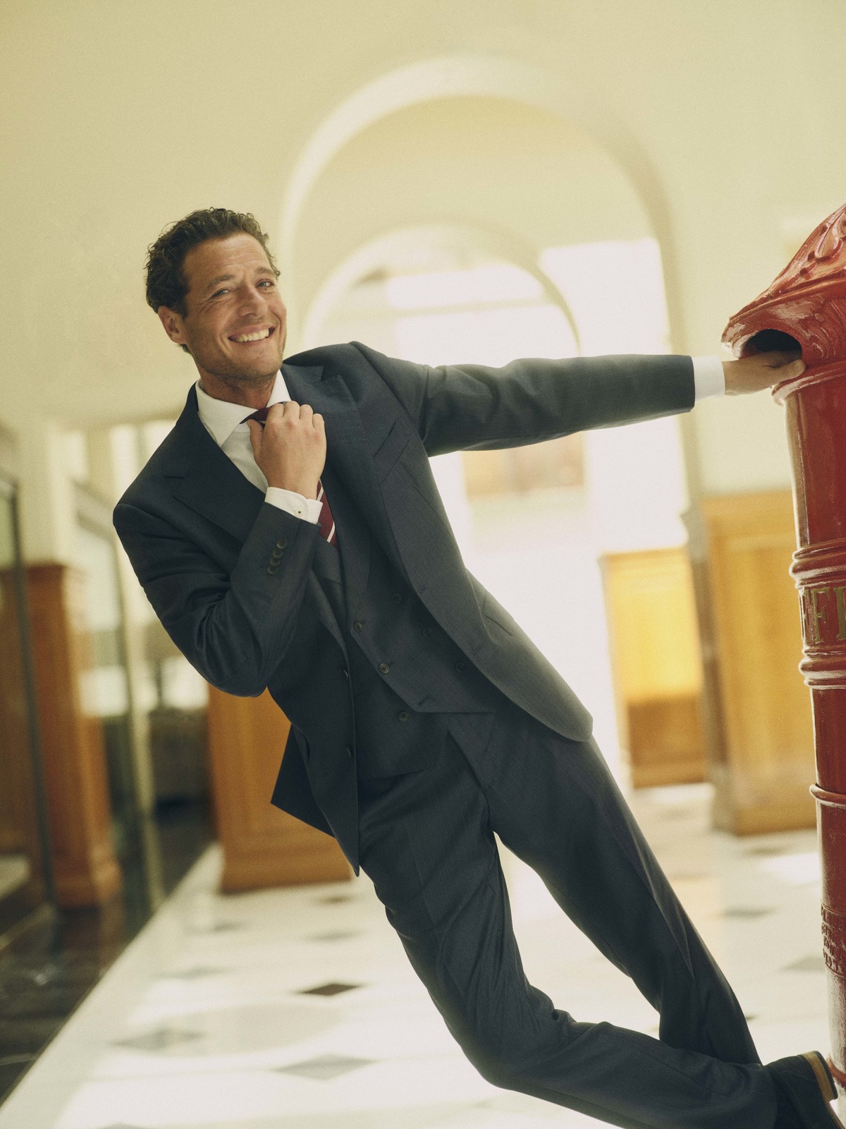 Smiling adult man in a suit leans against a red pillar indoors, adjusting his tie and looking towards the camera in a studio setting.