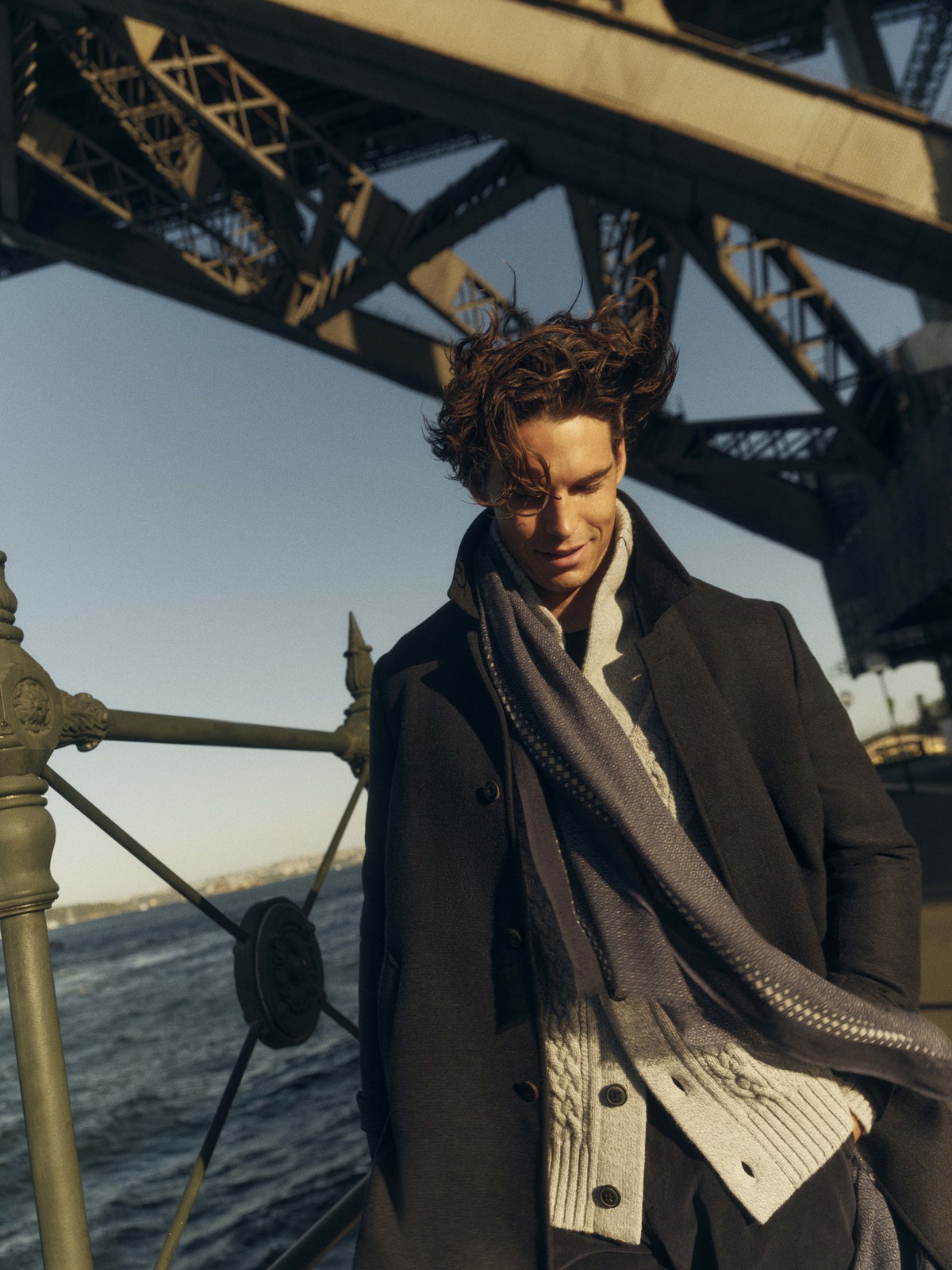 Adult male model in a black coat and grey scarf poses near a railing with a bridge in the background on an overcast day.