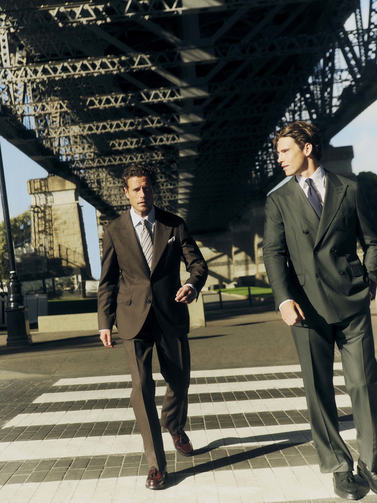 Two adult men in dark suits walking on a crosswalk with a metal bridge in the background on a bright, sunny day in an urban setting.