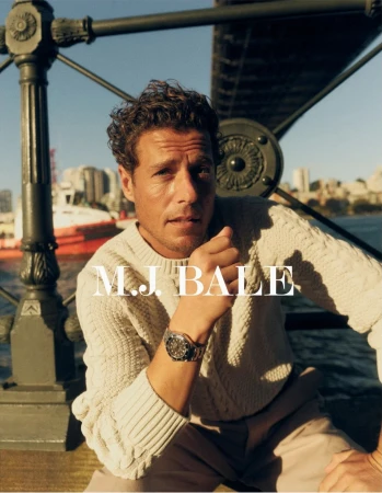 A man wearing a cream-colored sweater and a watch poses outdoors near a metal structure, with a cityscape and body of water visible.