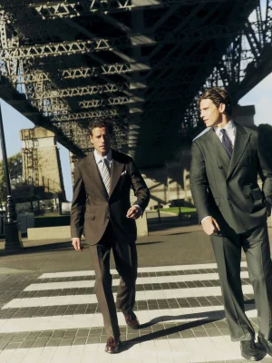 Two men in tailored suits walking across a paved crosswalk beneath a large metal bridge structure.