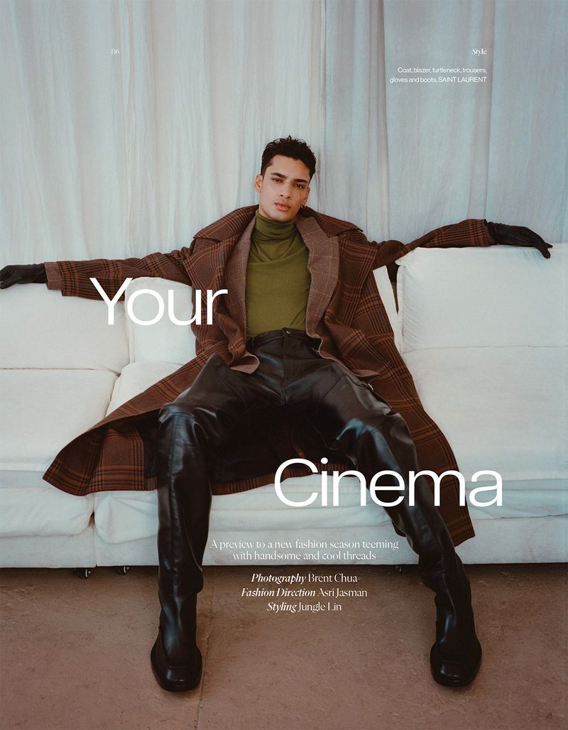 Fashion editorial shot of a male model in a brown coat and leather pants on a white sofa in a studio setting, showcasing Saint Laurent boots.