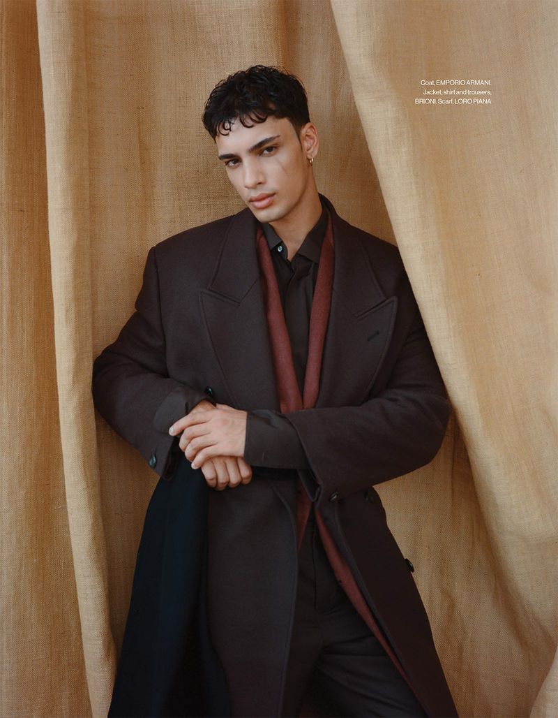 Fashionable young man poses indoors wearing a dark brown overcoat and a dark shirt with a tan canvas backdrop for a portrait shoot.
