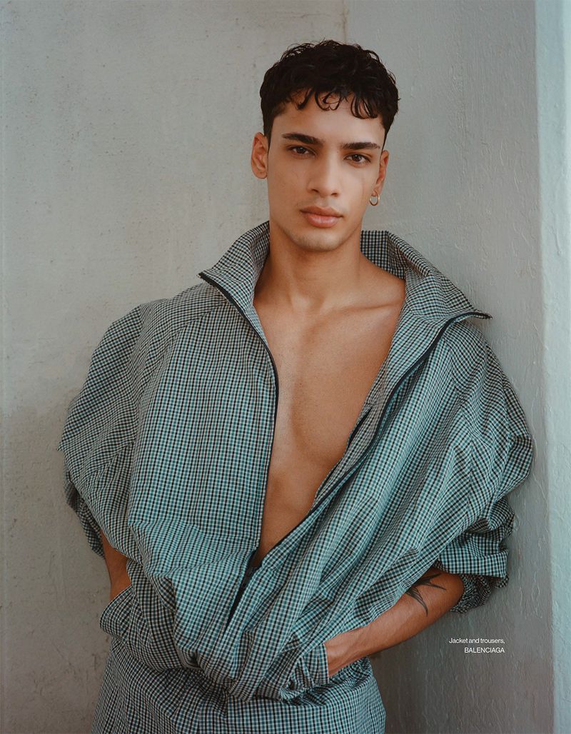 Fashion shot of a young adult male model posing indoors wearing a checkered jacket with a zipper and matching pants against a plain wall.