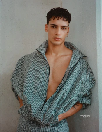 Fashion shot of a young adult male model posing indoors wearing a checkered jacket with a zipper and matching pants against a plain wall.