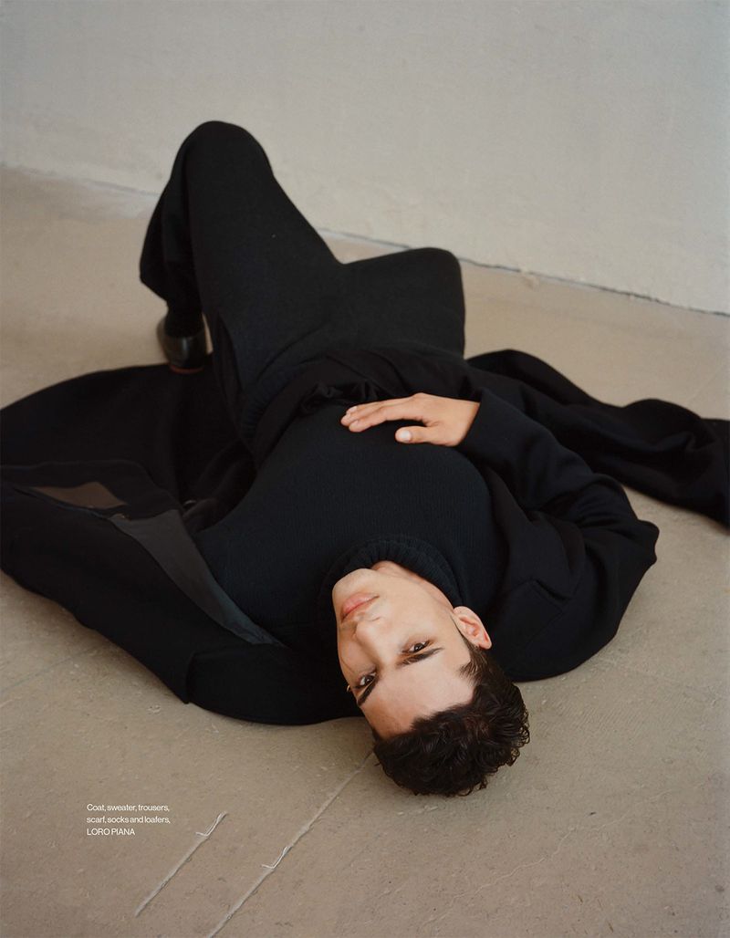 A young adult male lies on his back in a dark outfit against a neutral background, looking towards the viewer, with studio lighting.