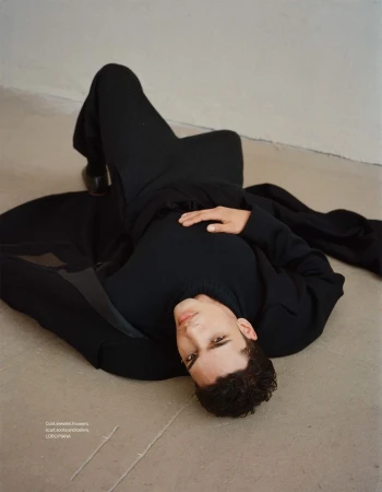 A young adult male lies on his back in a dark outfit against a neutral background, looking towards the viewer, with studio lighting.