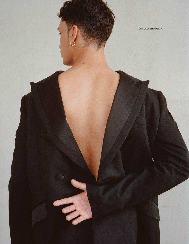 Fashion portrait of a back-facing adult male wearing a black, oversized tuxedo jacket with an open back, accessorized with gold earrings.