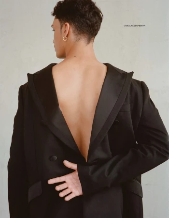 Fashion portrait of a back-facing adult male wearing a black, oversized tuxedo jacket with an open back, accessorized with gold earrings.