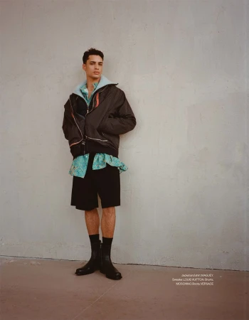 Model Mase Somanlall in a dark jacket, printed shirt, black shorts, and black leather boots against a neutral textured wall.