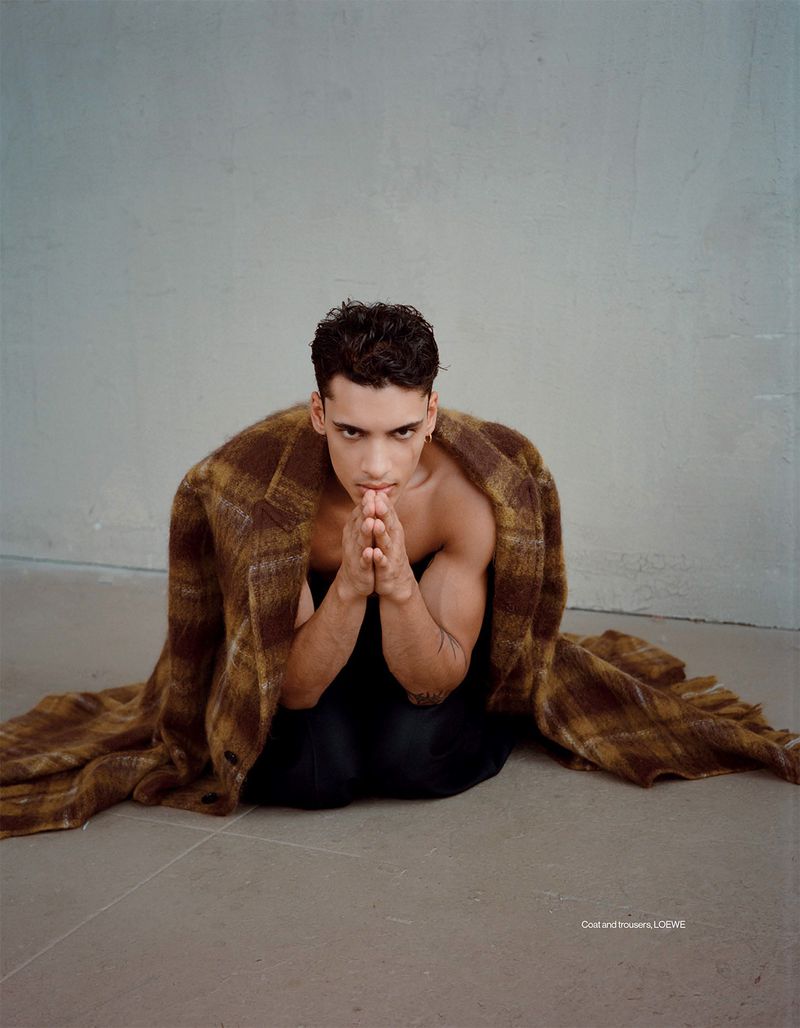 Man with muscular build in a kneeling position, draped with a brown plaid coat, hands together against a gray concrete backdrop.
