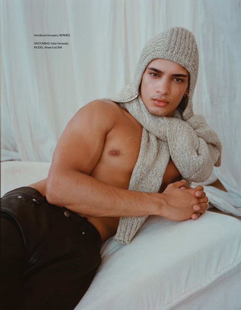 A tanned male model lies on a bed wearing a knit hat and scarf, showcasing fashion details in a studio with a draped backdrop.