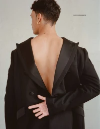 Rear view of a man with short dark hair wearing a reversed black tailored blazer exposing his bare back against a pale studio wall.