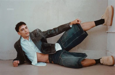 Male model lying on the floor in a dark leather jacket, white shirt, denim shorts, and slip-on shoes against a textured wall.