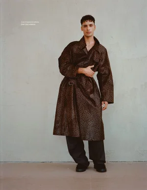 Full-length portrait of a male model wearing a patterned trench coat and dark trousers against a neutral wall.