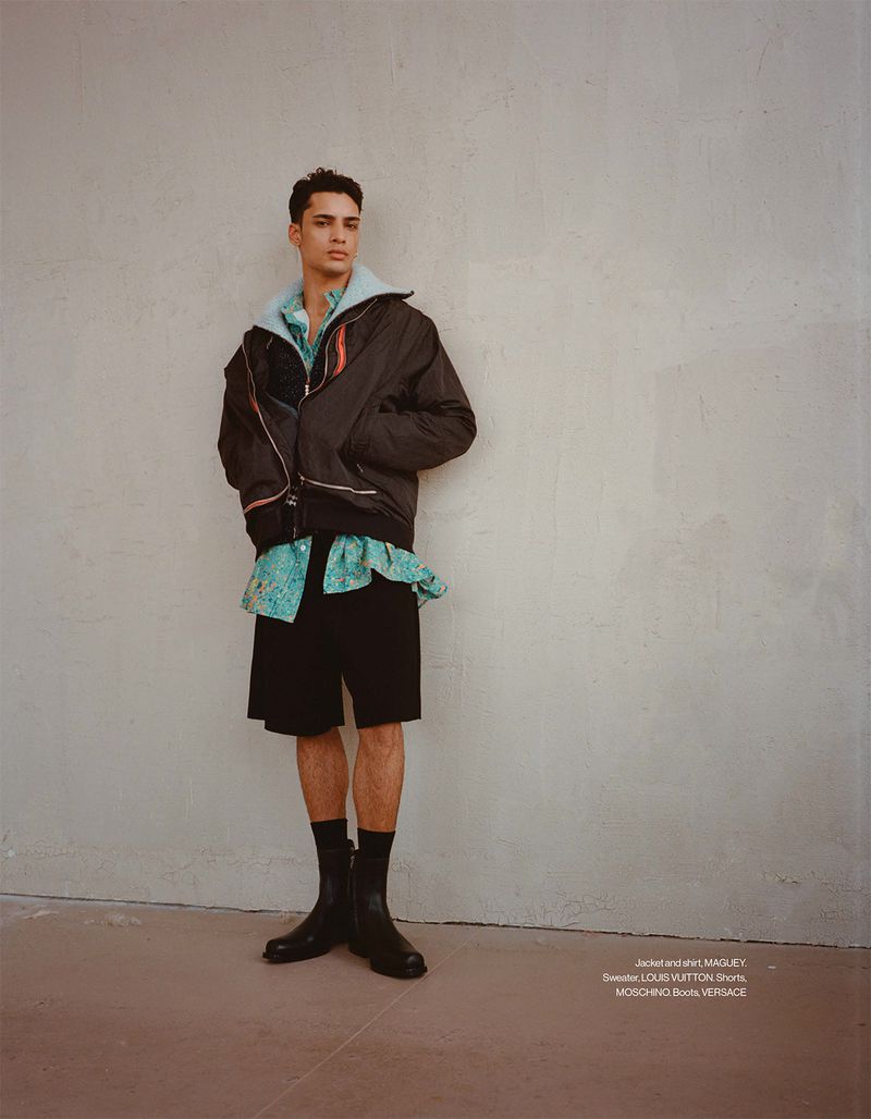 Model Mase Somanlall in a dark jacket, printed shirt, black shorts, and black leather boots against a neutral textured wall.