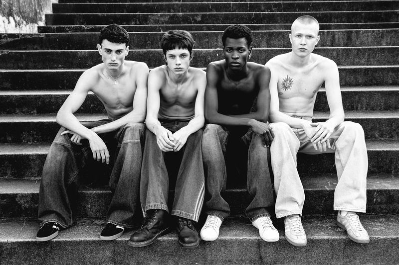 Four young adult men sit on outdoor stone steps in a black-and-white portrait. They wear casual pants and no shirts, set against stair background.