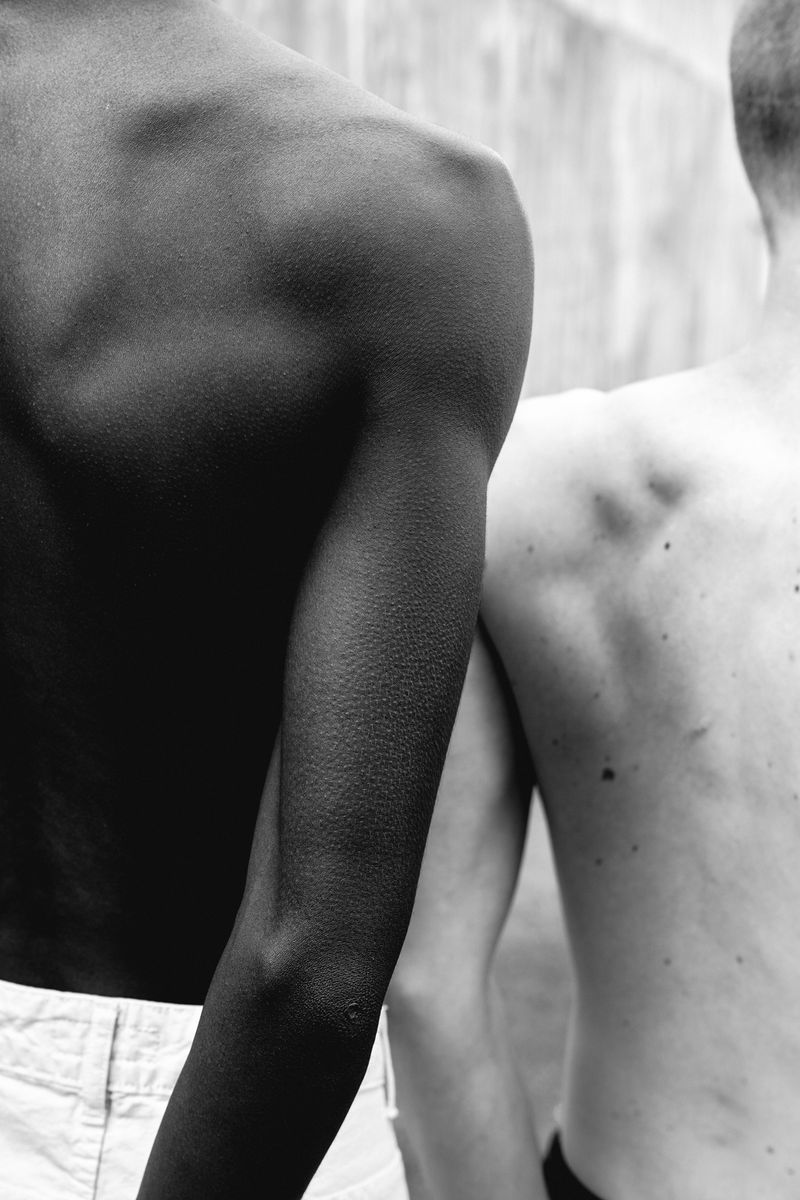 Back view of an African American adult and a Caucasian adult standing outdoors, captured in black and white. Focus on skin and shoulders.