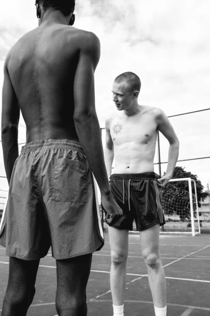 Monochrome shot of two shirtless men on an outdoor court. One man is facing away, while the other is looking at him with a slight smile.