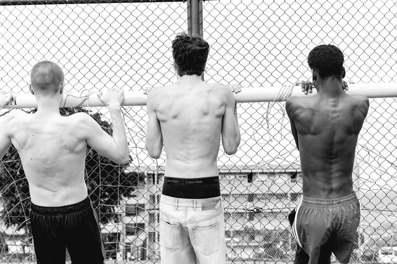 Three shirtless men stand at a chain-link fence in a black and white outdoor shot; one wears jeans, another running shorts.
