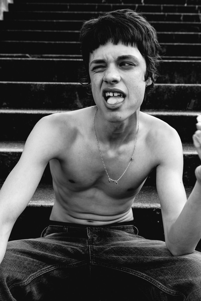 Black and white portrait of a young adult male sitting on concrete steps wearing denim jeans and a necklace, expressive face and bare chest.
