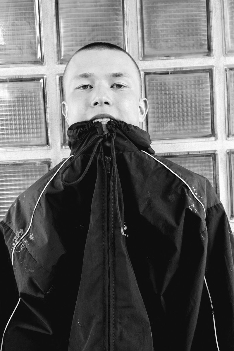 Portrait of an Asian adult male in a black hooded jacket, close-up shot against a windowed backdrop in a stark black and white aesthetic.