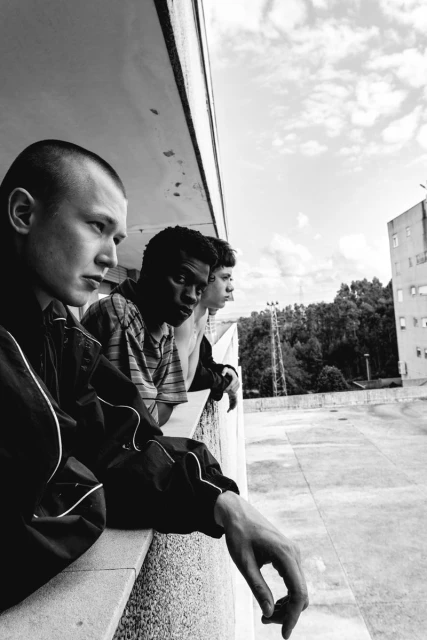 Black and white shot of three young men leaning against a ledge outdoors, looking out at an urban area with trees and buildings.