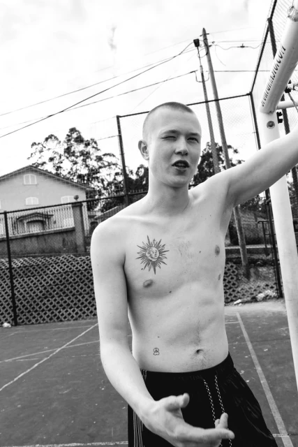 Monochrome portrait of a shirtless adult male with a shaved head, tattoo on chest, and standing outdoors on a sports court.