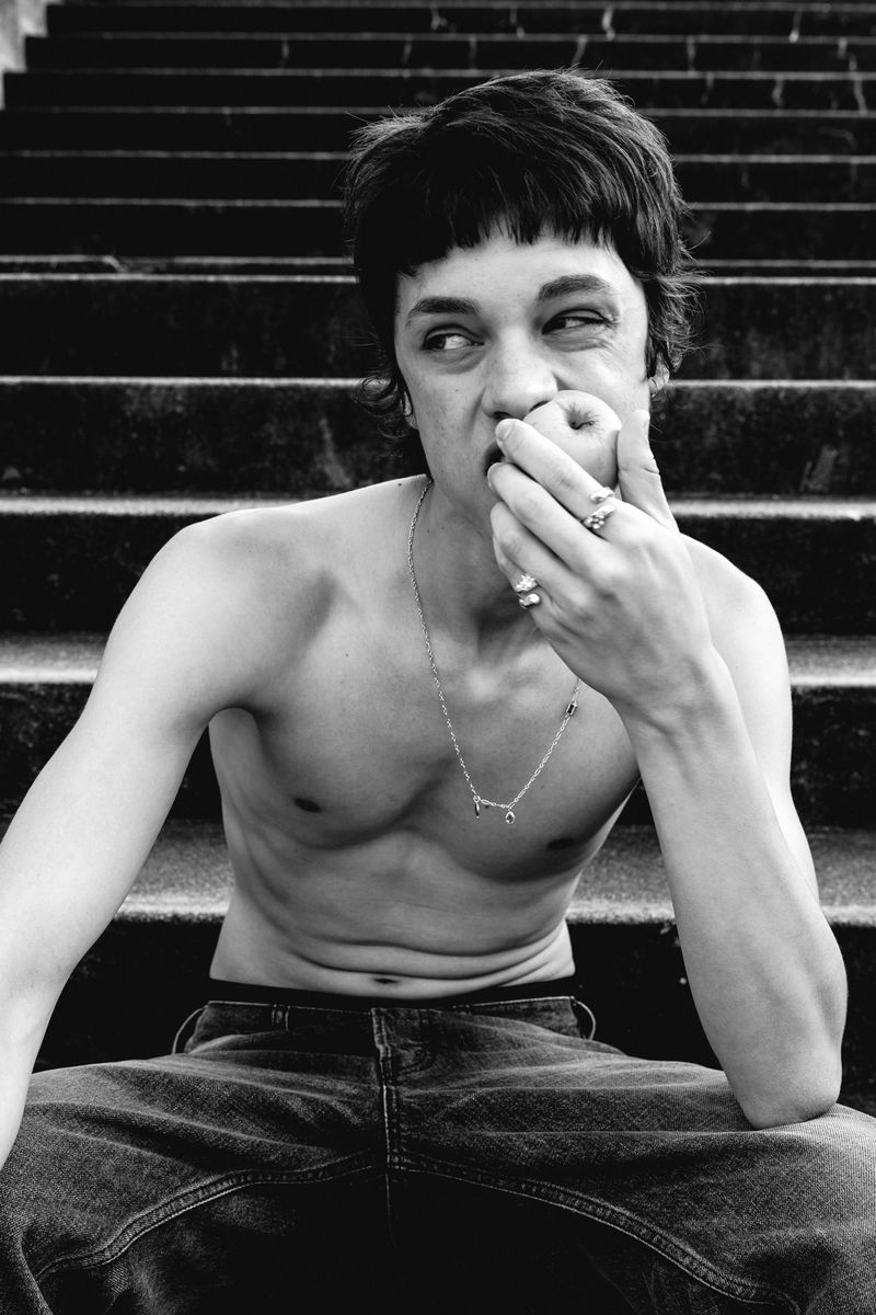 Black and white portrait of a shirtless young man sitting on outdoor steps while biting an apple, wearing jeans and silver jewellery.