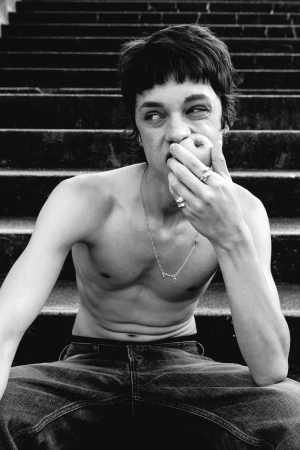 Black and white portrait of a shirtless young man sitting on outdoor steps while biting an apple, wearing jeans and silver jewellery.