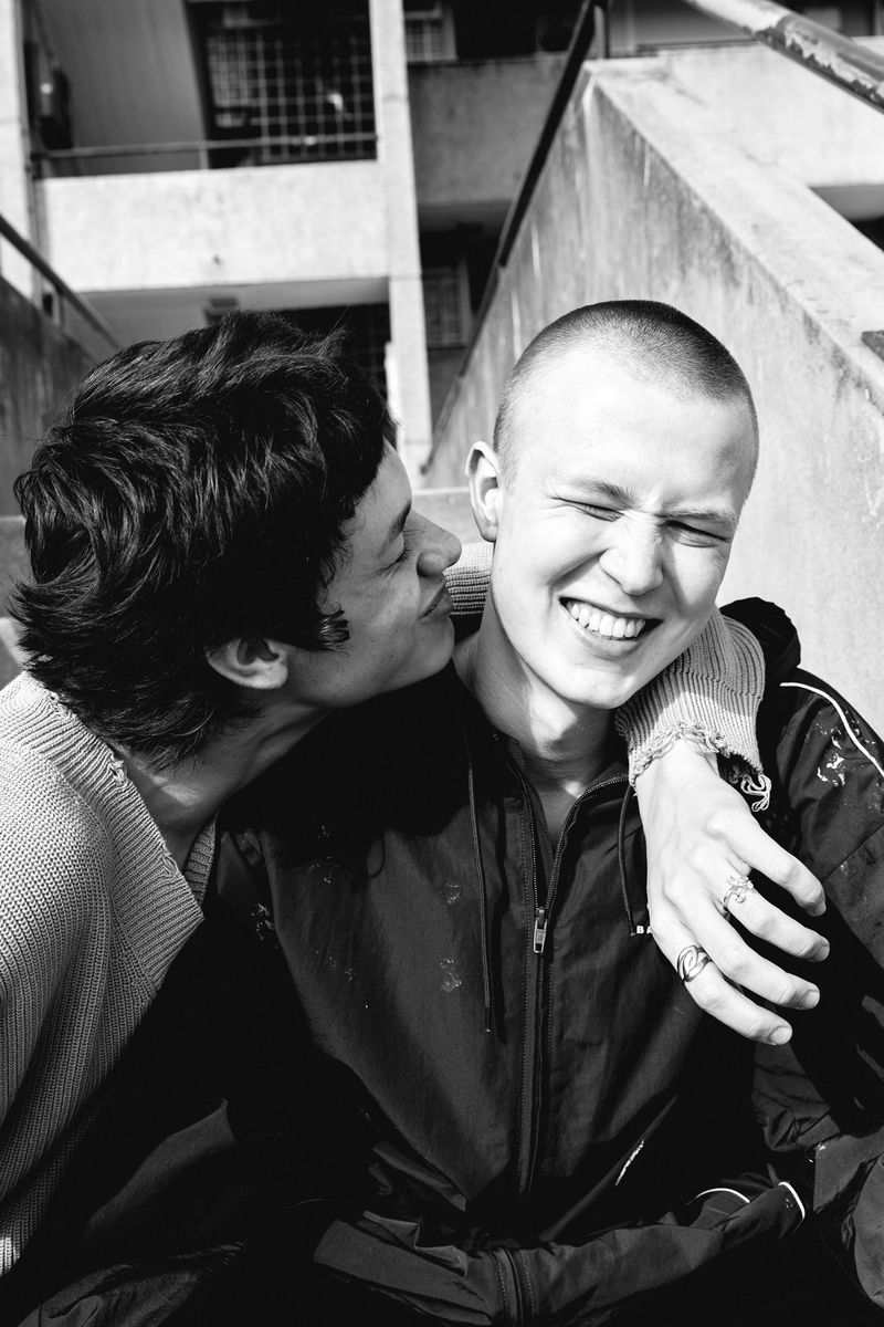 Black and white shot of two men embracing in urban setting with bright natural light. One man kisses the other's cheek.