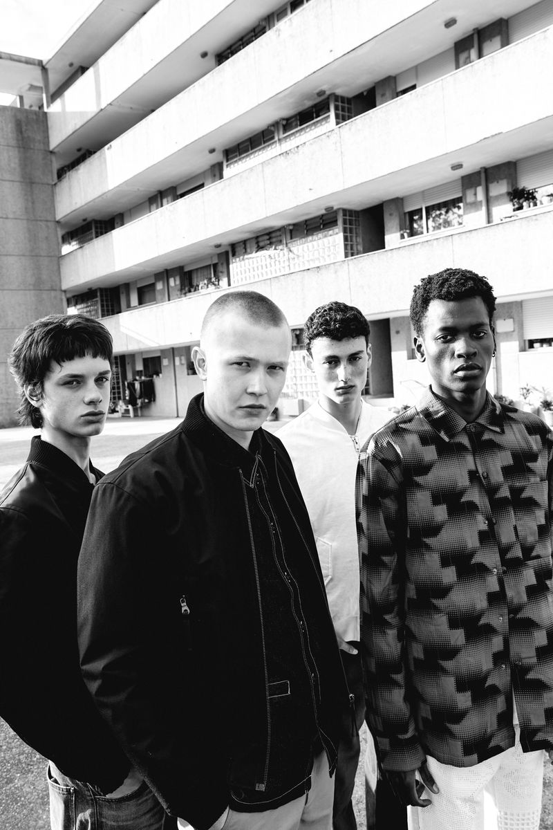 Black and white shot of four young adult men in casual fashion standing in front of a multi-story concrete building on a sunny day.