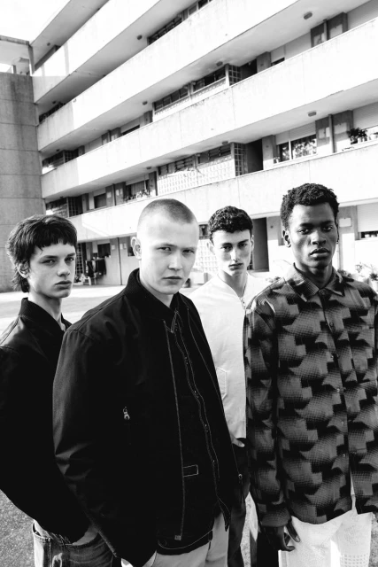 Black and white shot of four young adult men in casual fashion standing in front of a multi-story concrete building on a sunny day.