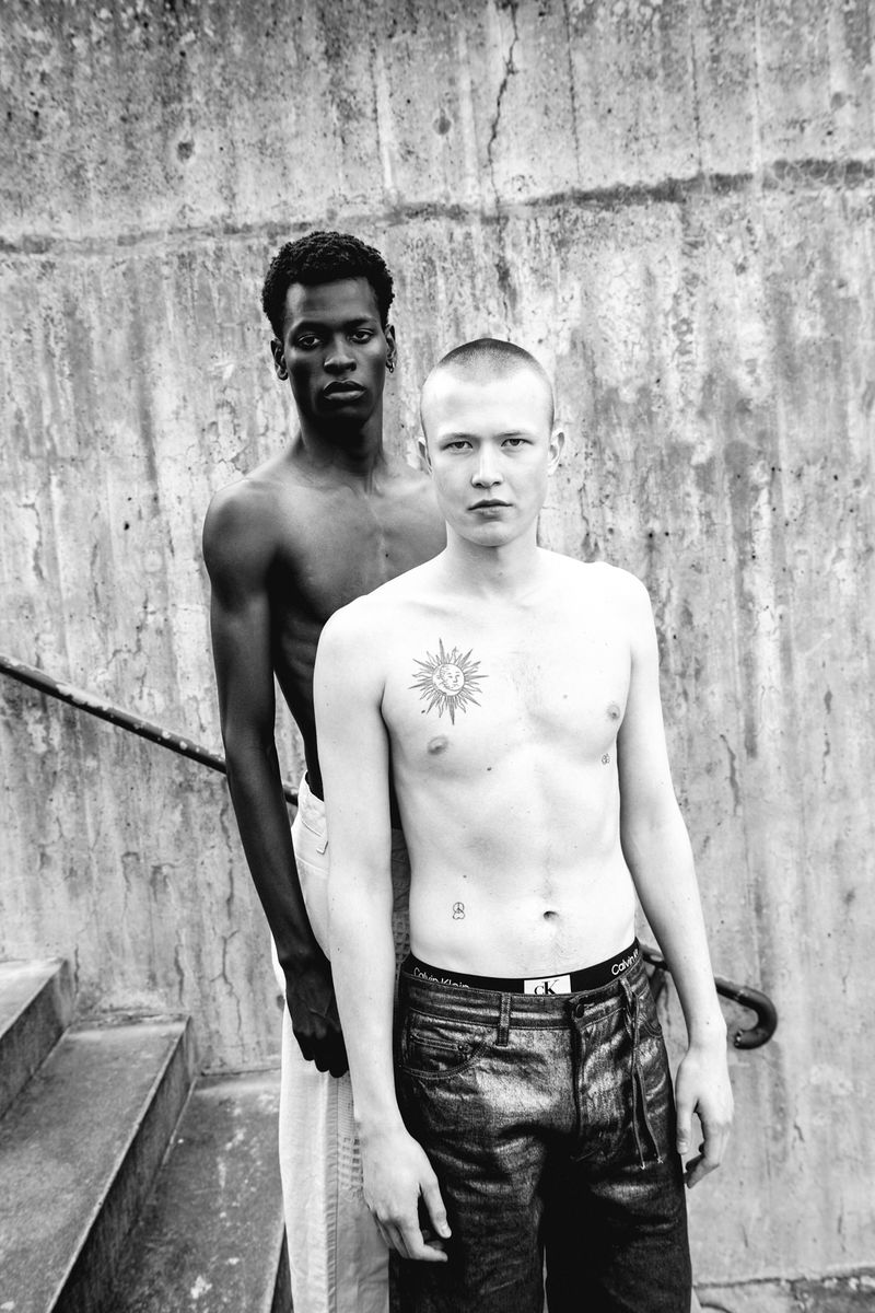 Black and white portrait of two shirtless men standing in front of a textured concrete wall and steps, with a serious expression.