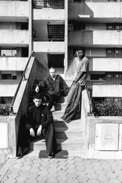 Three young men pose on concrete steps in front of a large building in black and white. Urban setting, casual clothing, direct gaze.