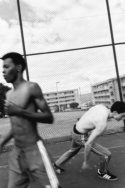 Two shirtless young men are captured in motion on an outdoor court, with a chain-link fence and residential buildings in the background.