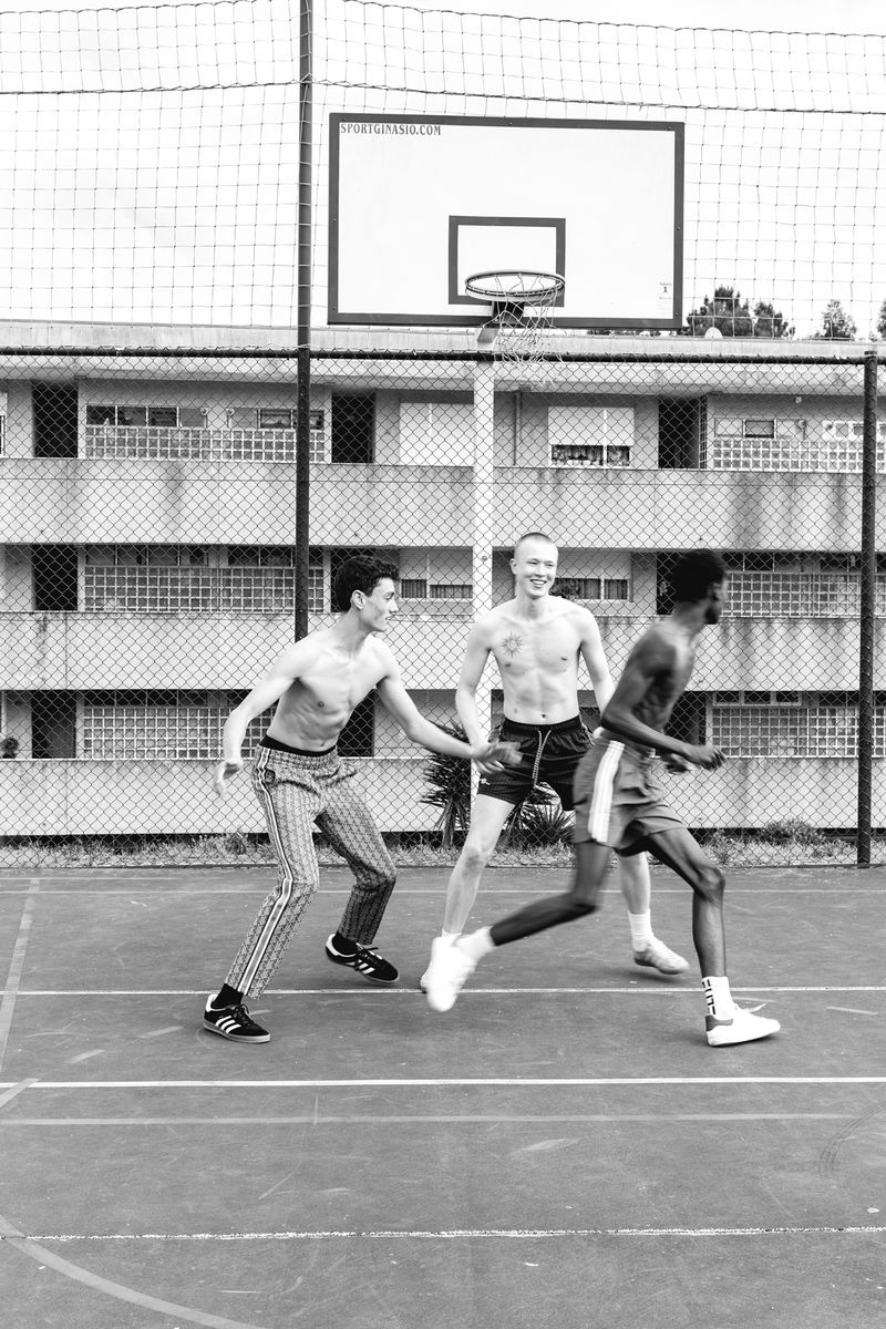 Three shirtless men play basketball on an outdoor court with a basketball hoop, surrounded by an apartment building, in black and white.