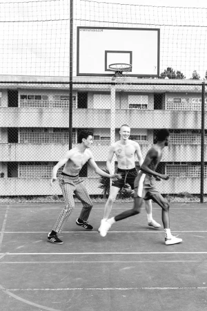 Three shirtless men play basketball on an outdoor court with a basketball hoop, surrounded by an apartment building, in black and white.