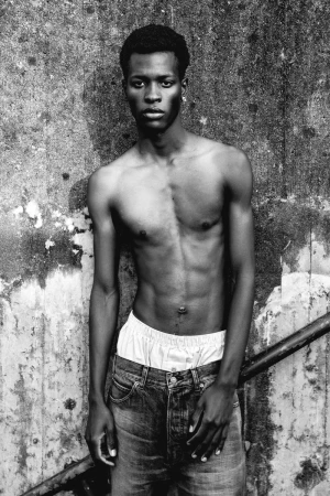 Black and white portrait of a young, muscular Black man in jeans, standing against a textured wall, looking directly at the camera.