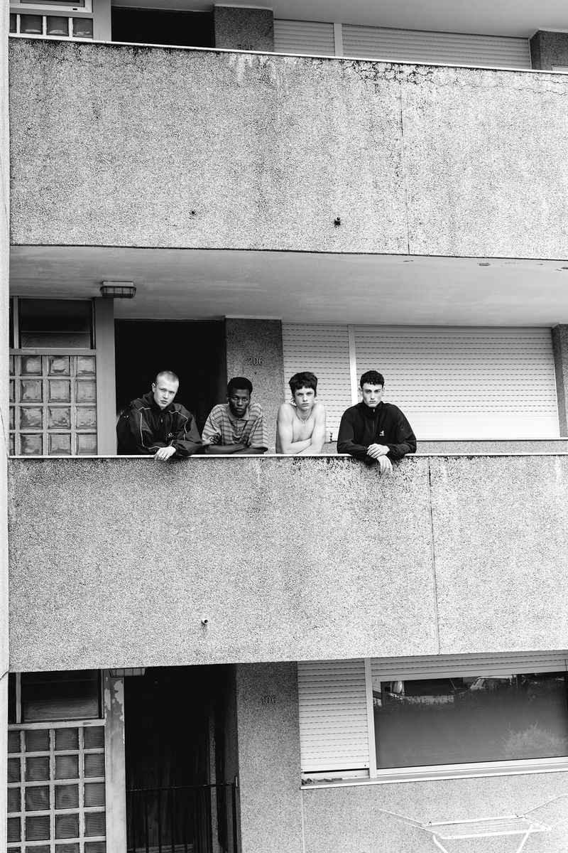 Four men stand on a concrete balcony overlooking the street. The monochrome photo features a residential building and a view of the outdoors.