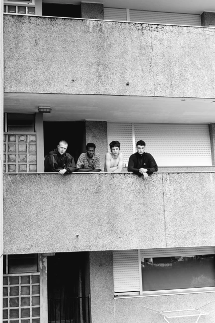 Four men stand on a concrete balcony overlooking the street. The monochrome photo features a residential building and a view of the outdoors.