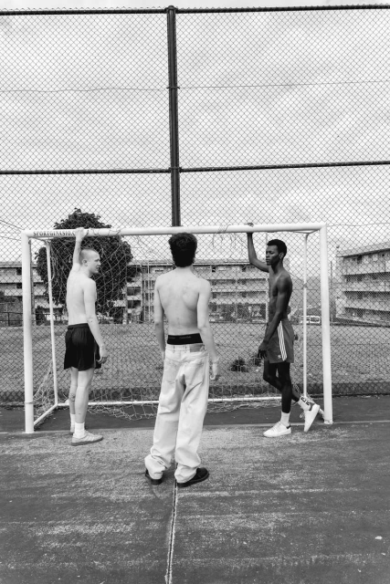 Three shirtless young men stand by a soccer goal on a concrete court, with apartment buildings and a chain-link fence in the background.