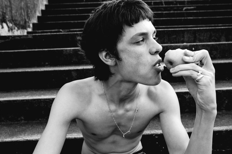 Monochrome portrait of shirtless young adult eating fruit outdoors on concrete steps, wearing necklace and rings, pensive expression.