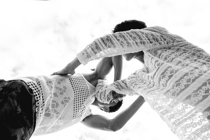 Two people in white lace tops are close together in a low-angle shot against a bright, overcast sky, creating a high-contrast black-and-white effect.