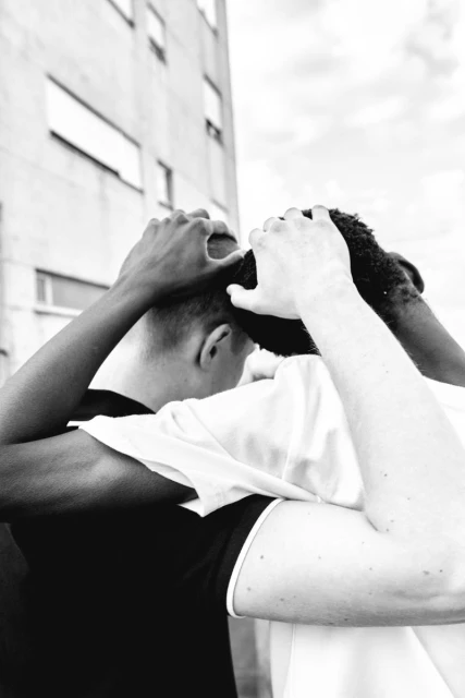 Black and white image of two young men embracing, one with dark skin and wearing a black shirt, the other with light skin, white shirt, outside.