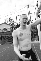 Black and white portrait of a shirtless young man with a chest tattoo on an outdoor court for Numero Netherlands.