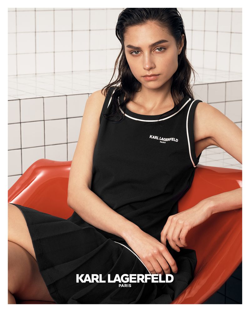 A woman models a black sleeveless dress with white trim detail in front of a tiled wall, seated in a bright orange chair, and logo text.