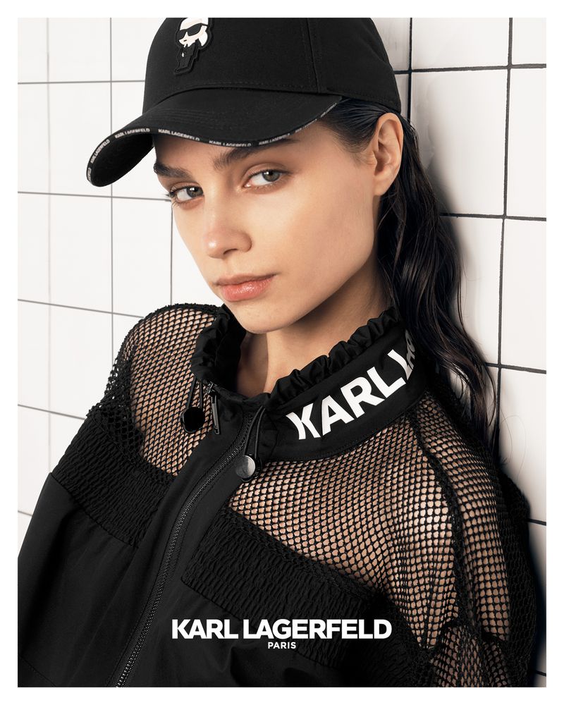 Model Ana Rossolovich in a black Karl Lagerfeld cap and mesh-panel jacket leaning against a white tiled wall.